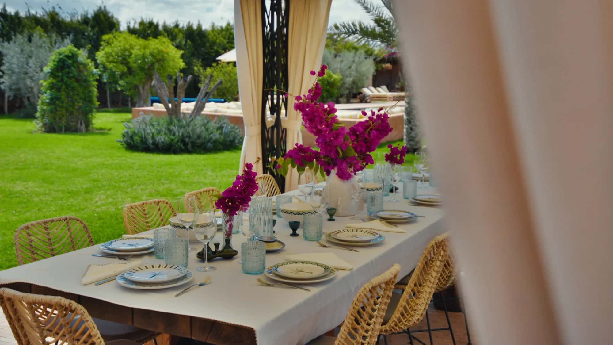 Outdoor dining table at Villa Yemna in Marrakech set for a meal, with fuchsia bougainvillea, rattan chairs and garden view.