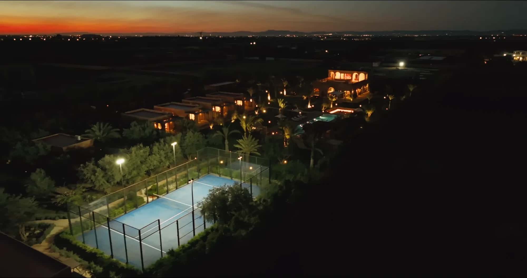 Twilight drone view of Villa Yemna in Marrakech with illuminated padel court, pool and orange horizon line over the plain.