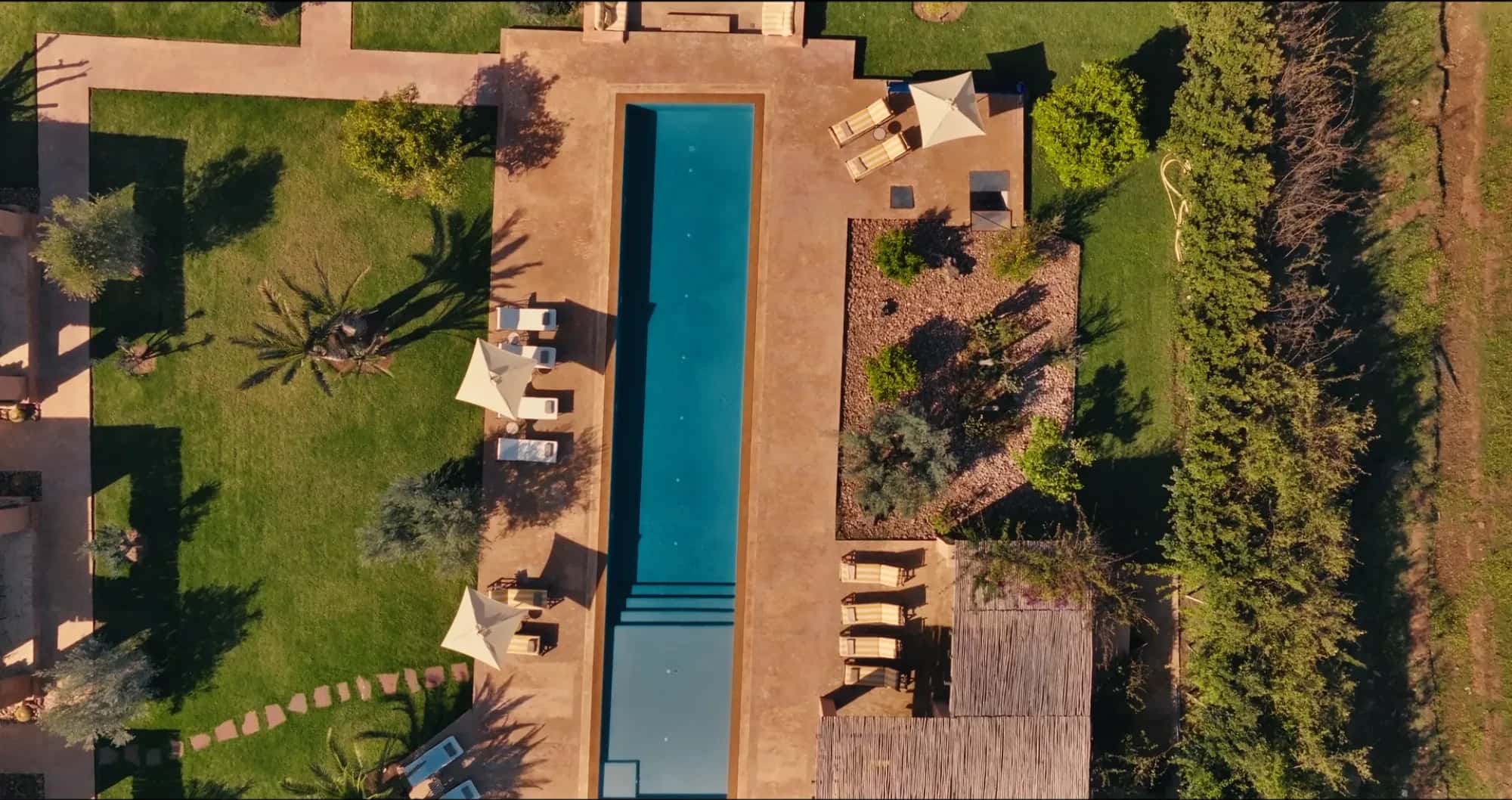 Aerial drone view of the 21m saltwater pool at Villa Yemna in Marrakech, with sun loungers, tadelakt deck and surrounding olive grove.
