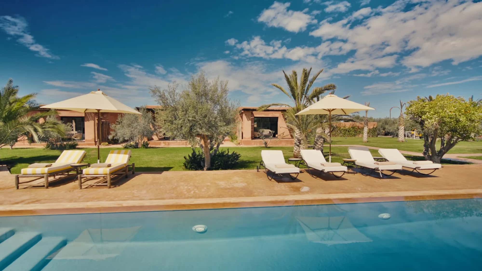 Pool deck at Villa Yemna in Marrakech with sun loungers, parasols, olive trees and a suite bungalow in the background.