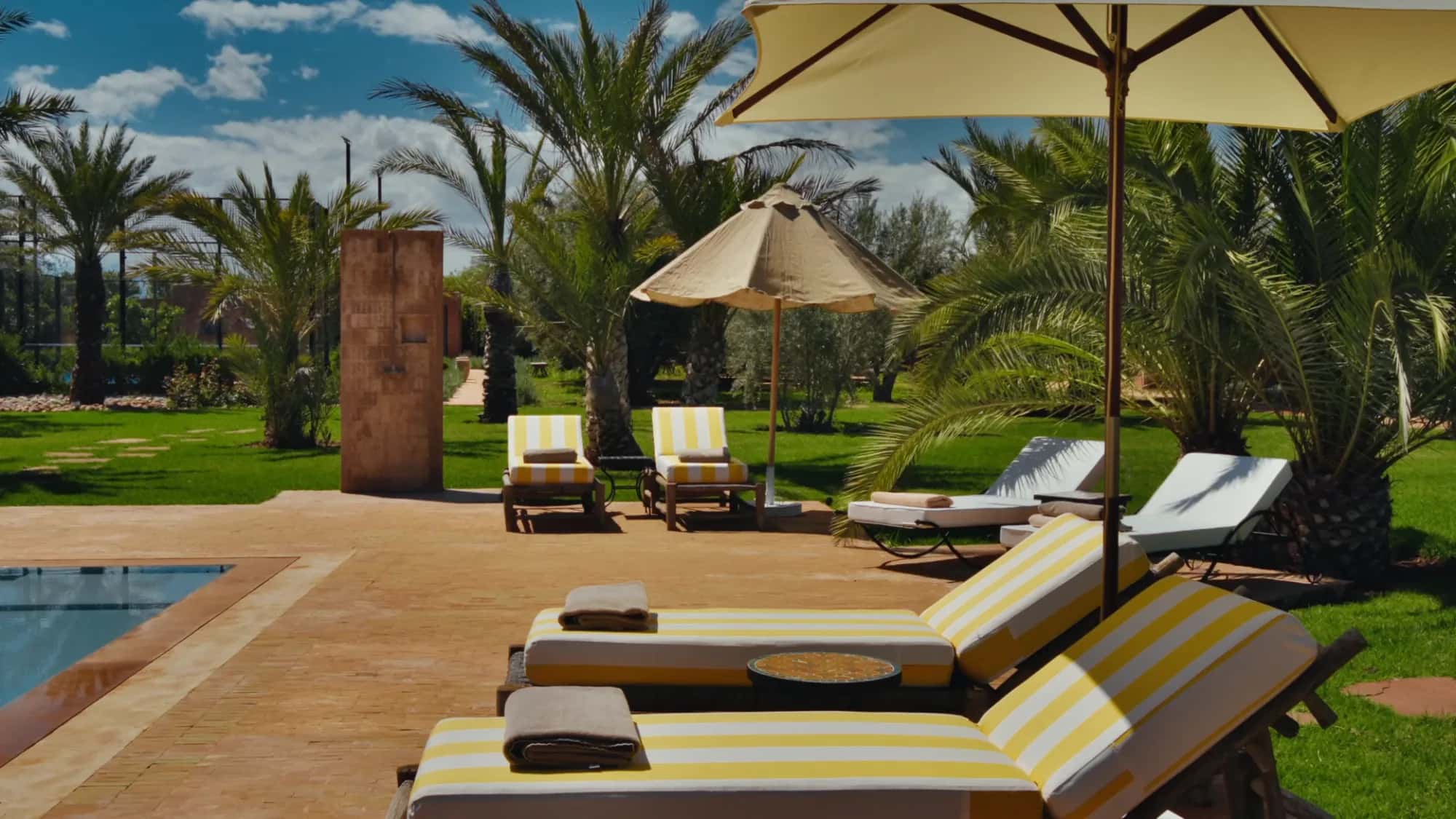 Yellow striped sun loungers by the pool at Villa Yemna in Marrakech, under parasols and date palm trees.