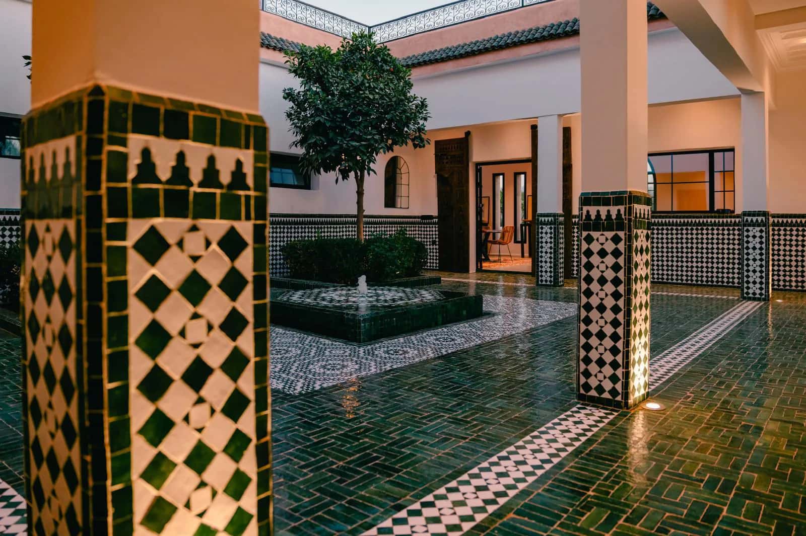 Villa Yemna riad courtyard at dusk, with glossy green zellige flooring, patterned columns, and warm interior lights.
