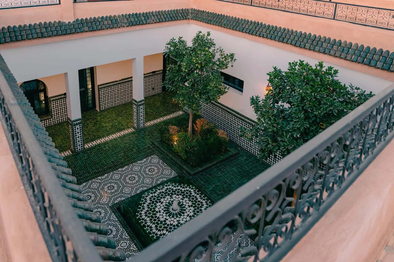 Elevated view of the Villa Yemna riad courtyard in Marrakech, with green zellige tiles, orange trees, a central fountain, and Moroccan arcades.