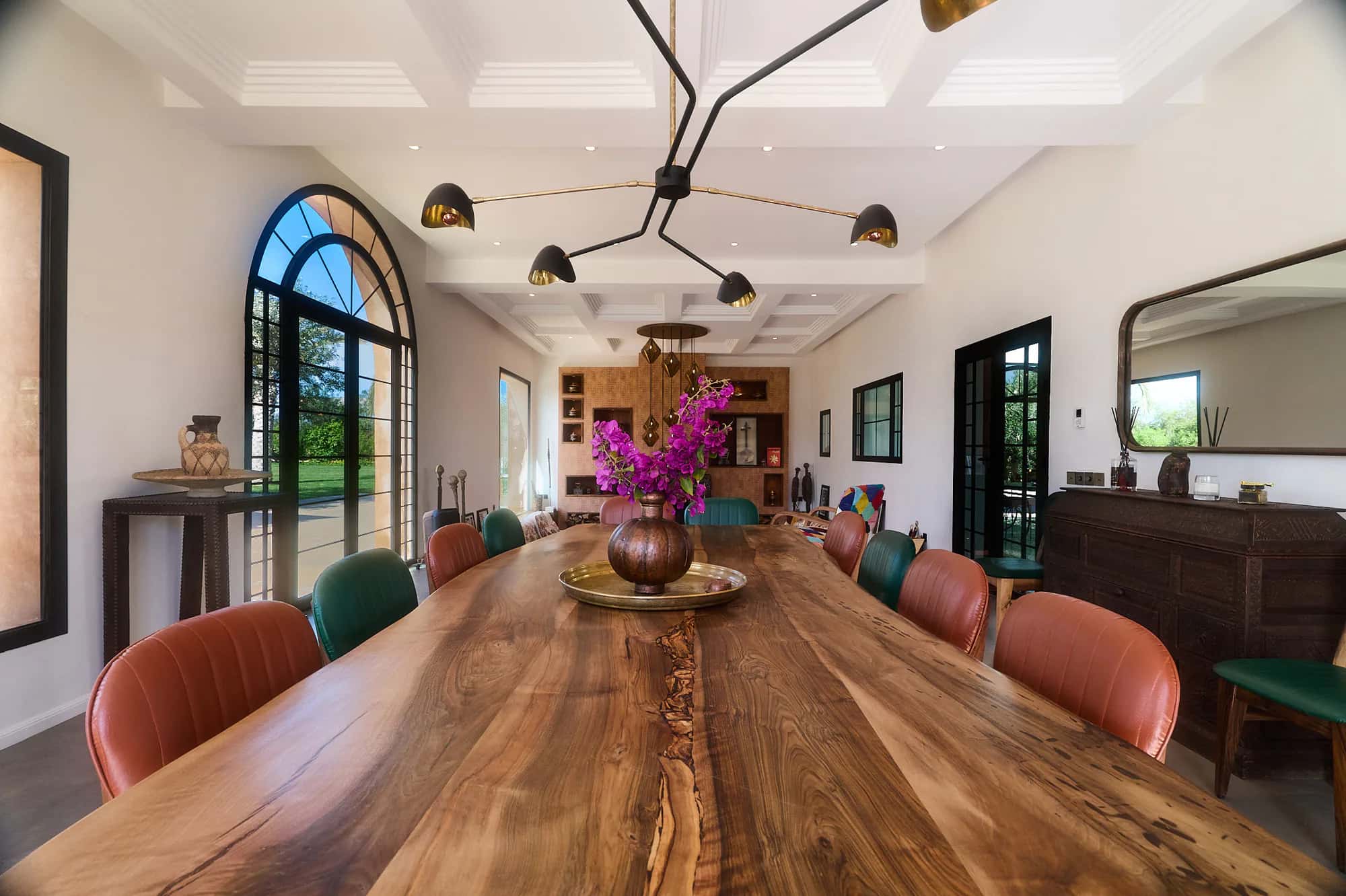 Villa Yemna riad dining room with a long solid wood table, colorful leather chairs, bougainvillea flowers, and Moroccan pendant lights.
