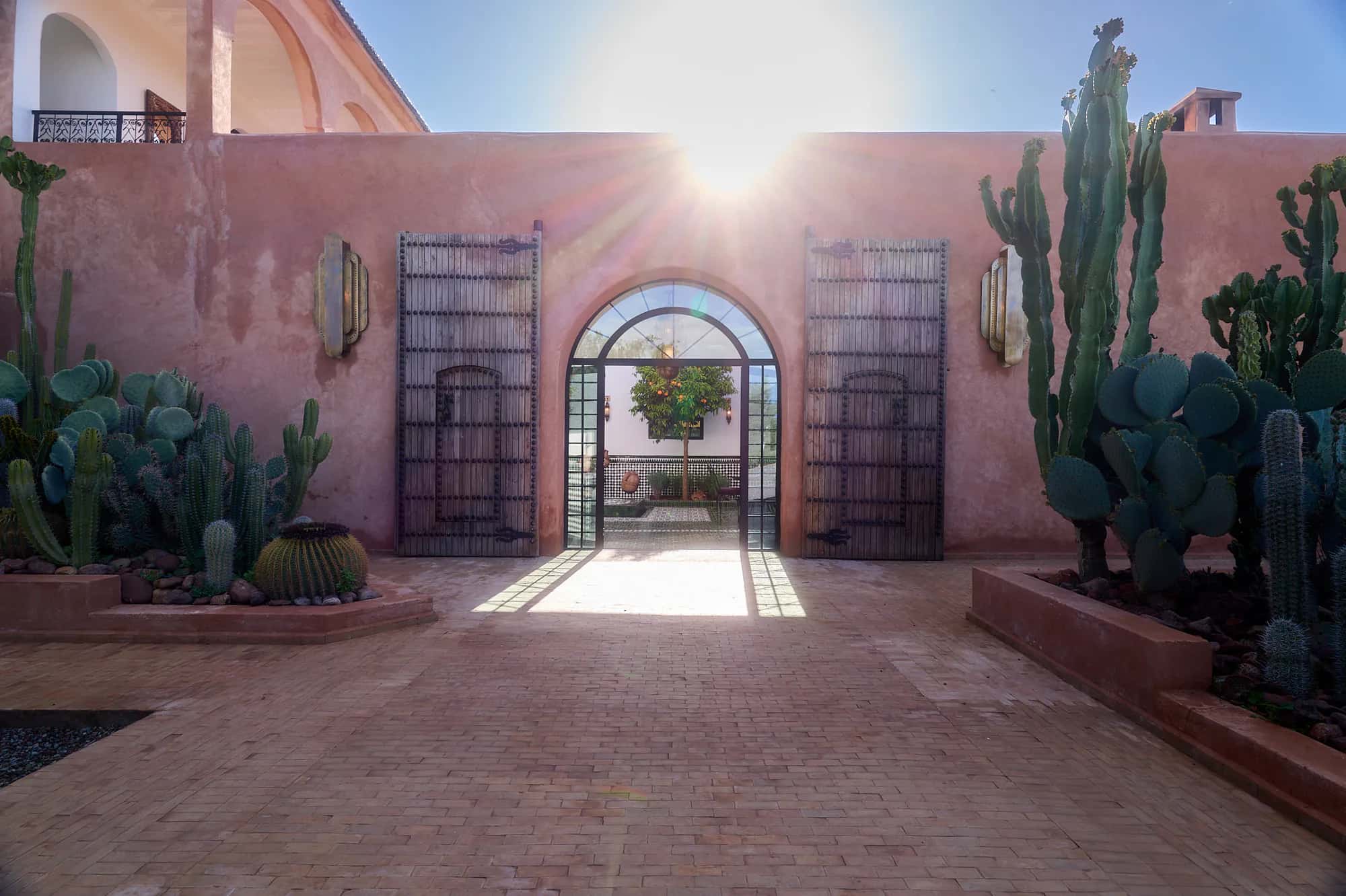 Sunlit entrance of Villa Yemna in Marrakech, with a cactus garden, large traditional wooden doors, and a view into the riad courtyard.