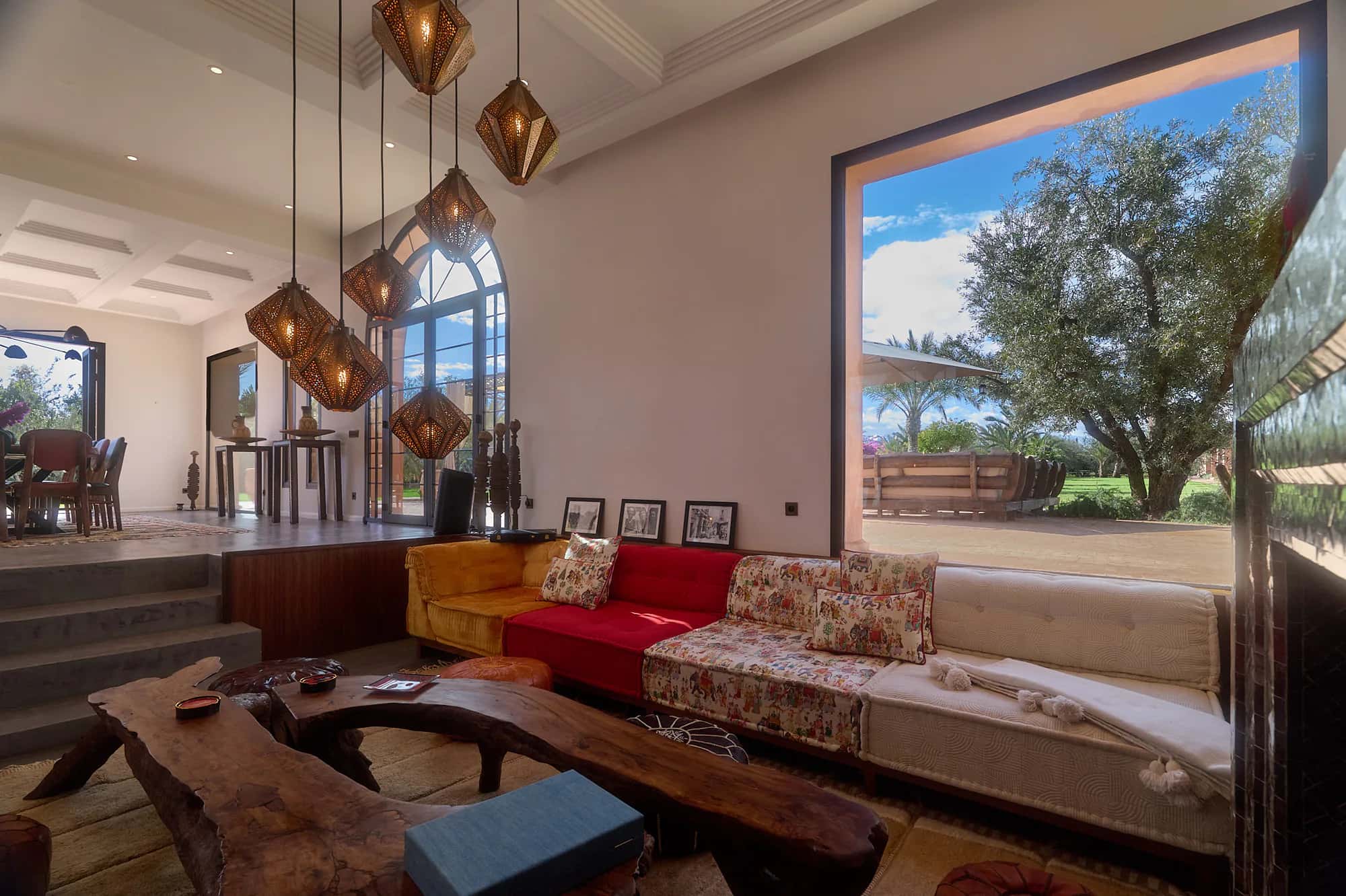Villa Yemna riad lounge with patterned sofas, pendant lanterns, a zellige fireplace, and a large window overlooking the garden.