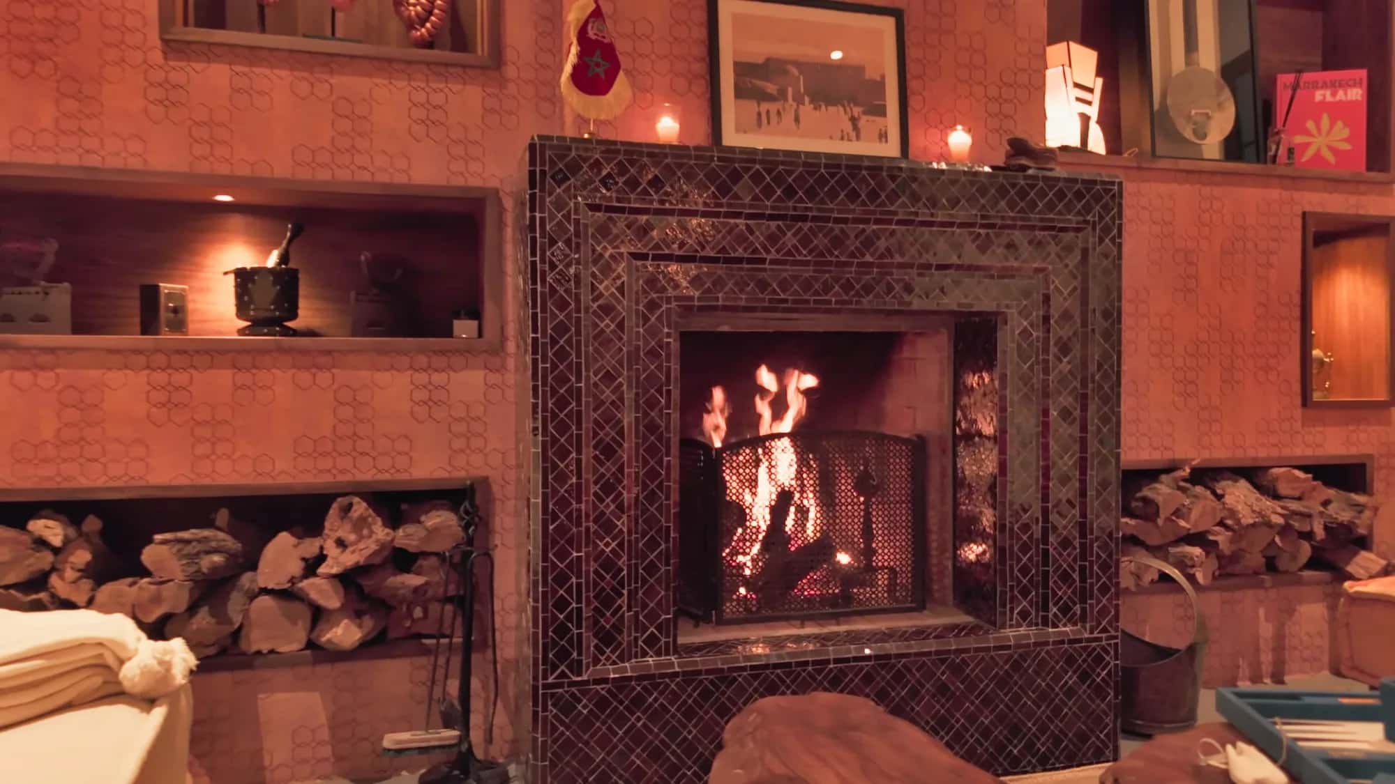Black zellige fireplace with crackling fire and stacked logs, in the Villa Yemna riad lounge in Marrakech.