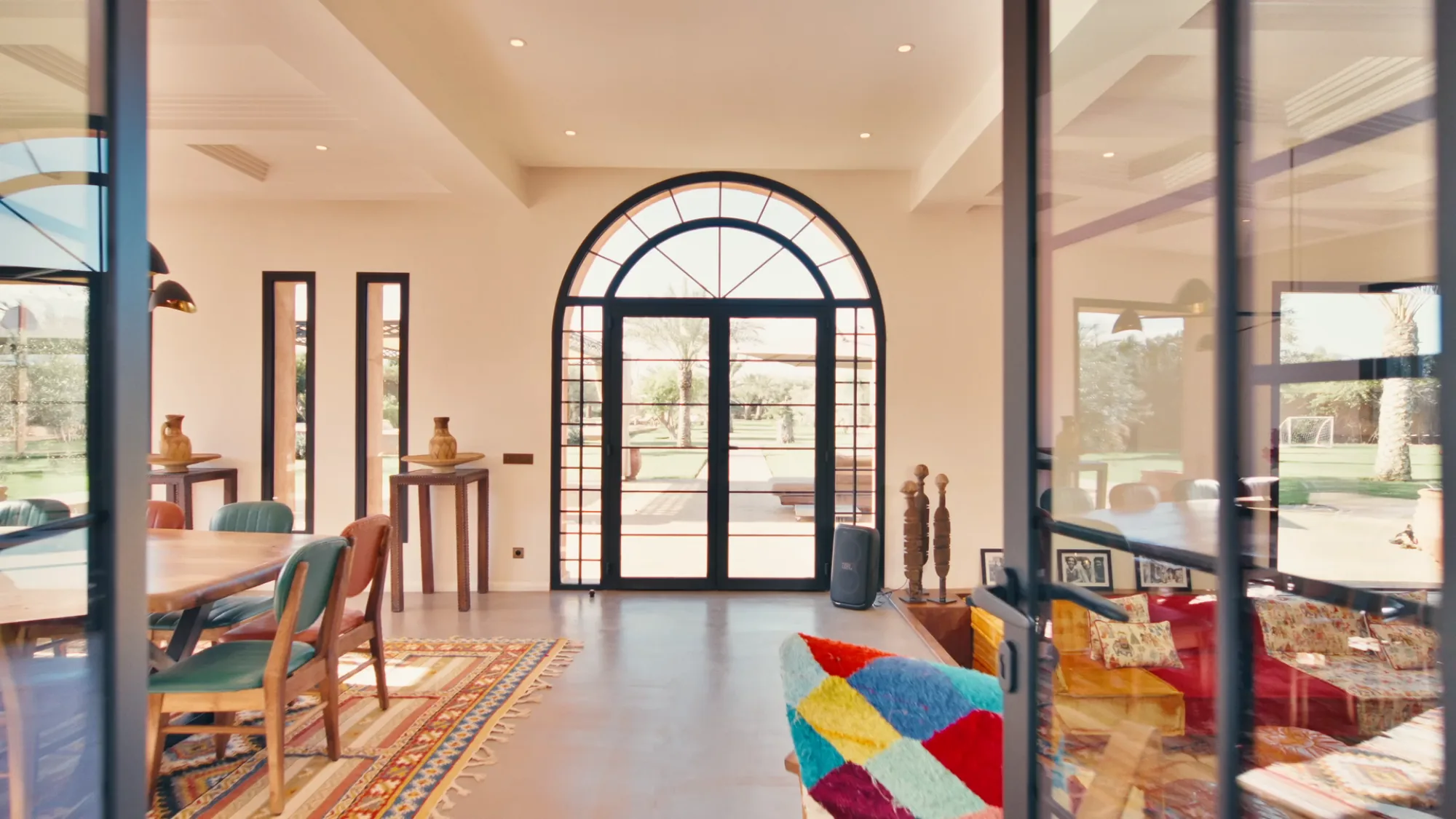 Villa Yemna riad living room in Marrakech with large arched window, multicolored patchwork sofa, Berber rug and African sculptures.