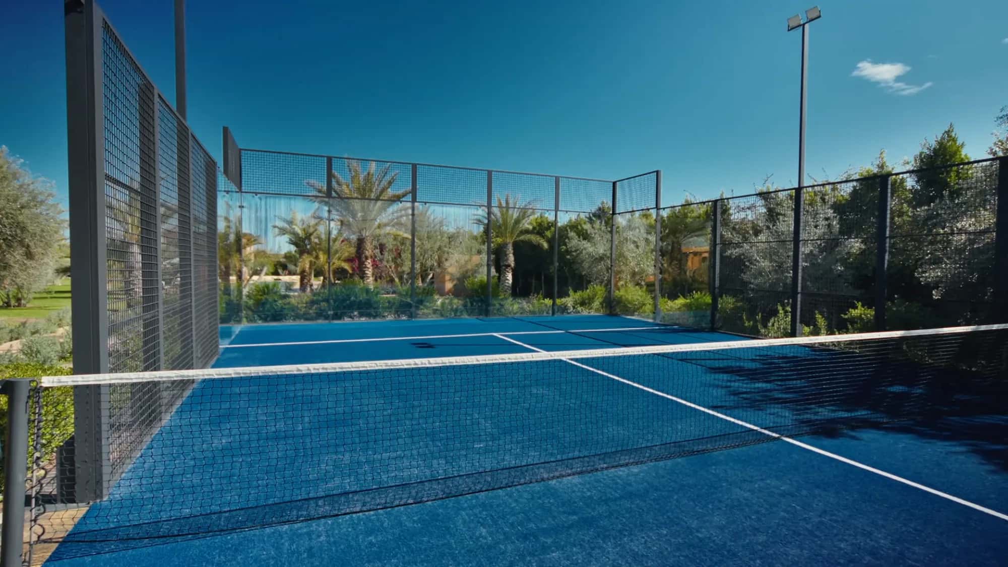 Private padel court at Villa Yemna in Marrakech viewed from inside, with blue playing surface, net and surrounding olive grove.