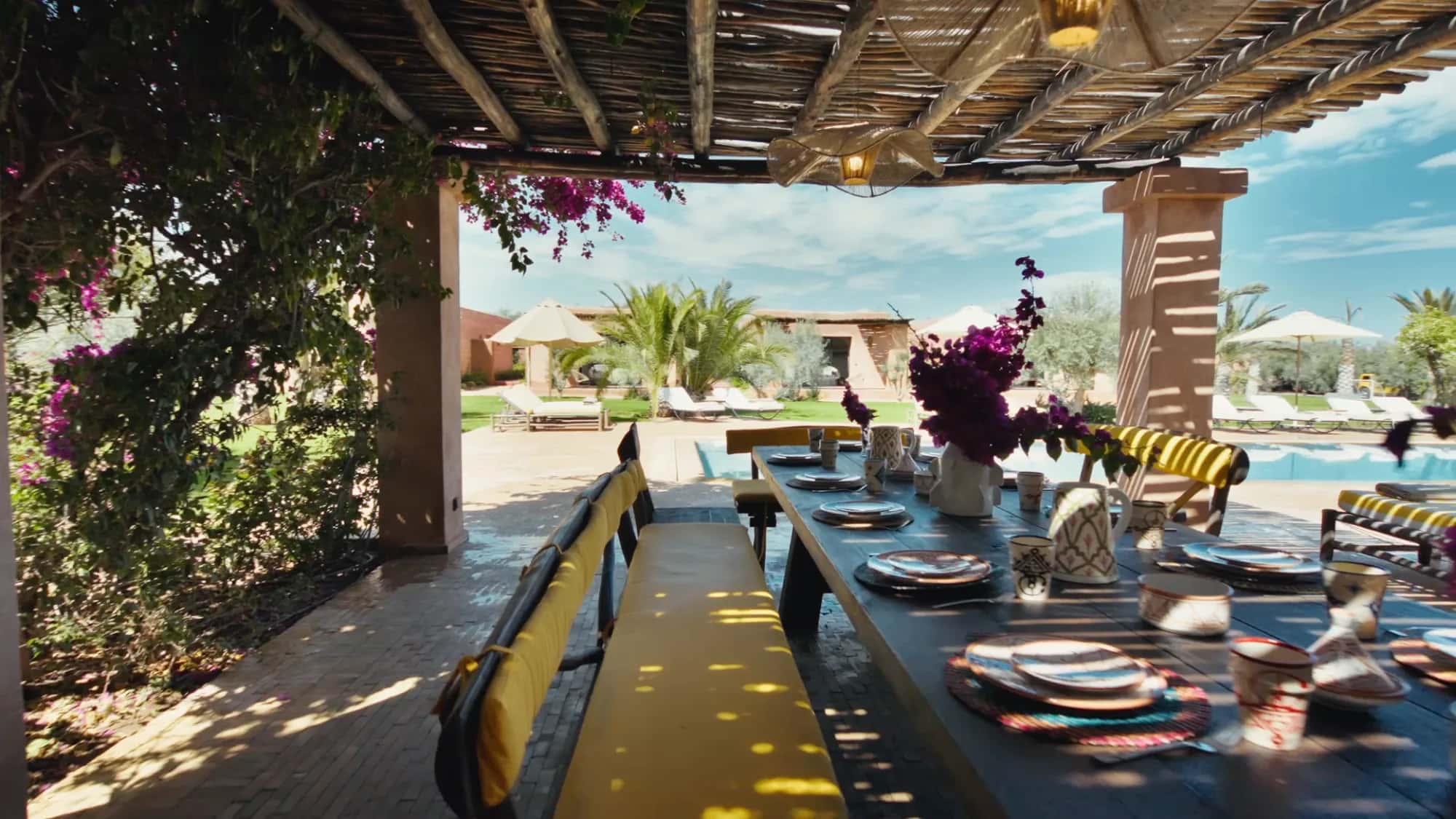 Poolside pergola dining table at Villa Yemna in Marrakech with striped cushions, bougainvillea and garden views.
