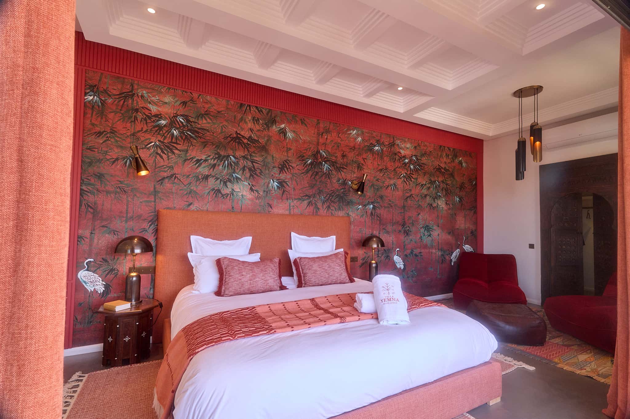 Suite Oukaimeden bedroom at Villa Yemna, with white bedding, terracotta cushions, a red armchair, and bamboo wall decor.