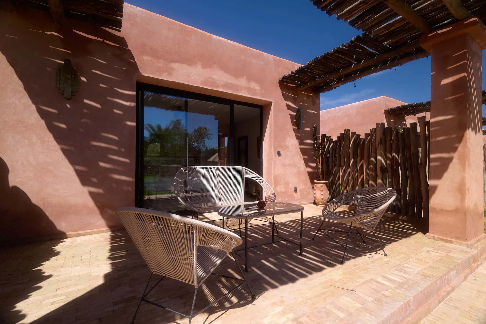 Private terrace of a Villa Yemna bungalow suite with woven chairs, a coffee table, pergola shadows, and terracotta walls.