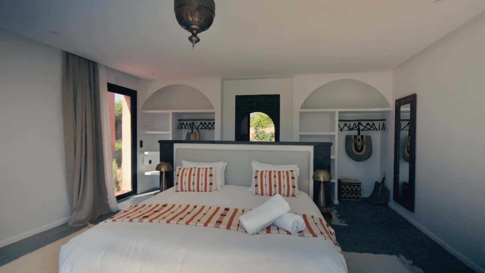 Villa Yemna suite bedroom in Marrakech with king-size bed in white linen, arched niches, terracotta striped pillows and Moroccan pendant light.