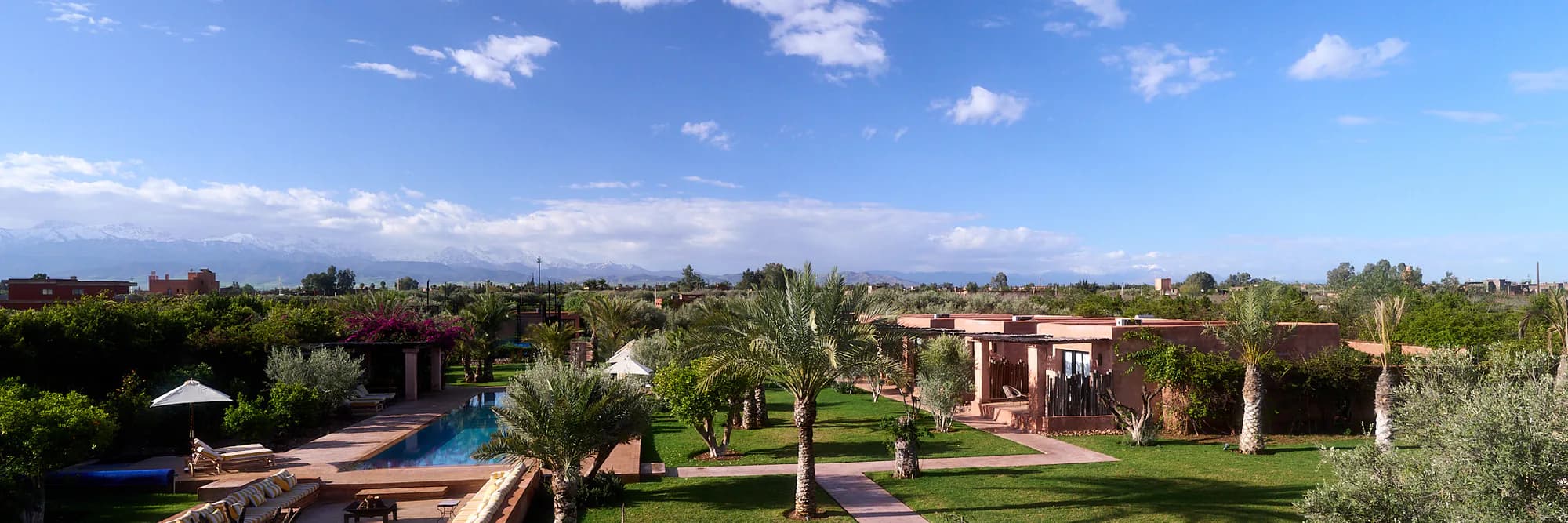 Panoramic view of the Villa Yemna estate with pool, gardens, palm trees, bungalow suites, and the Atlas Mountains.