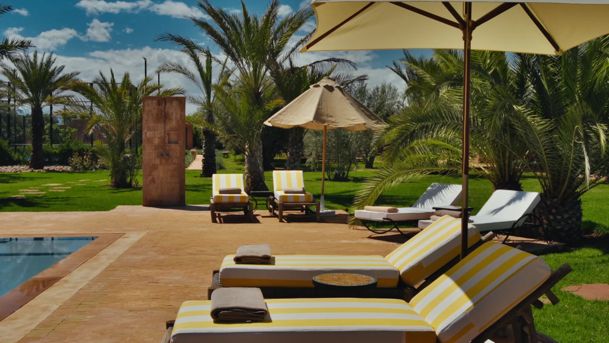 Transats à rayures jaunes au bord de la piscine de Villa Yemna à Marrakech, sous parasols et palmiers dattiers.