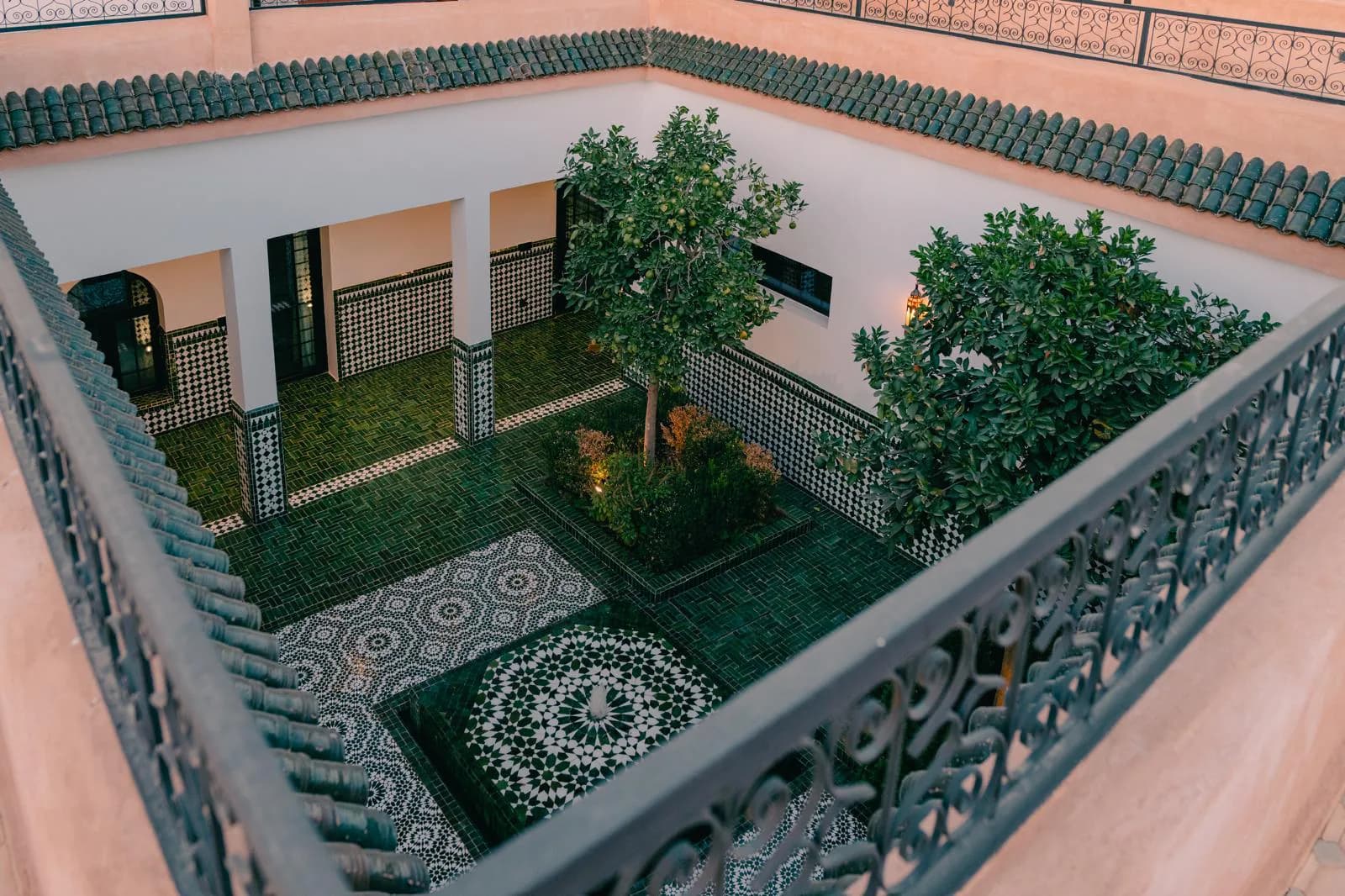 Elevated view of the Villa Yemna riad courtyard in Marrakech, with green zellige tiles, orange trees, a central fountain, and Moroccan arcades.