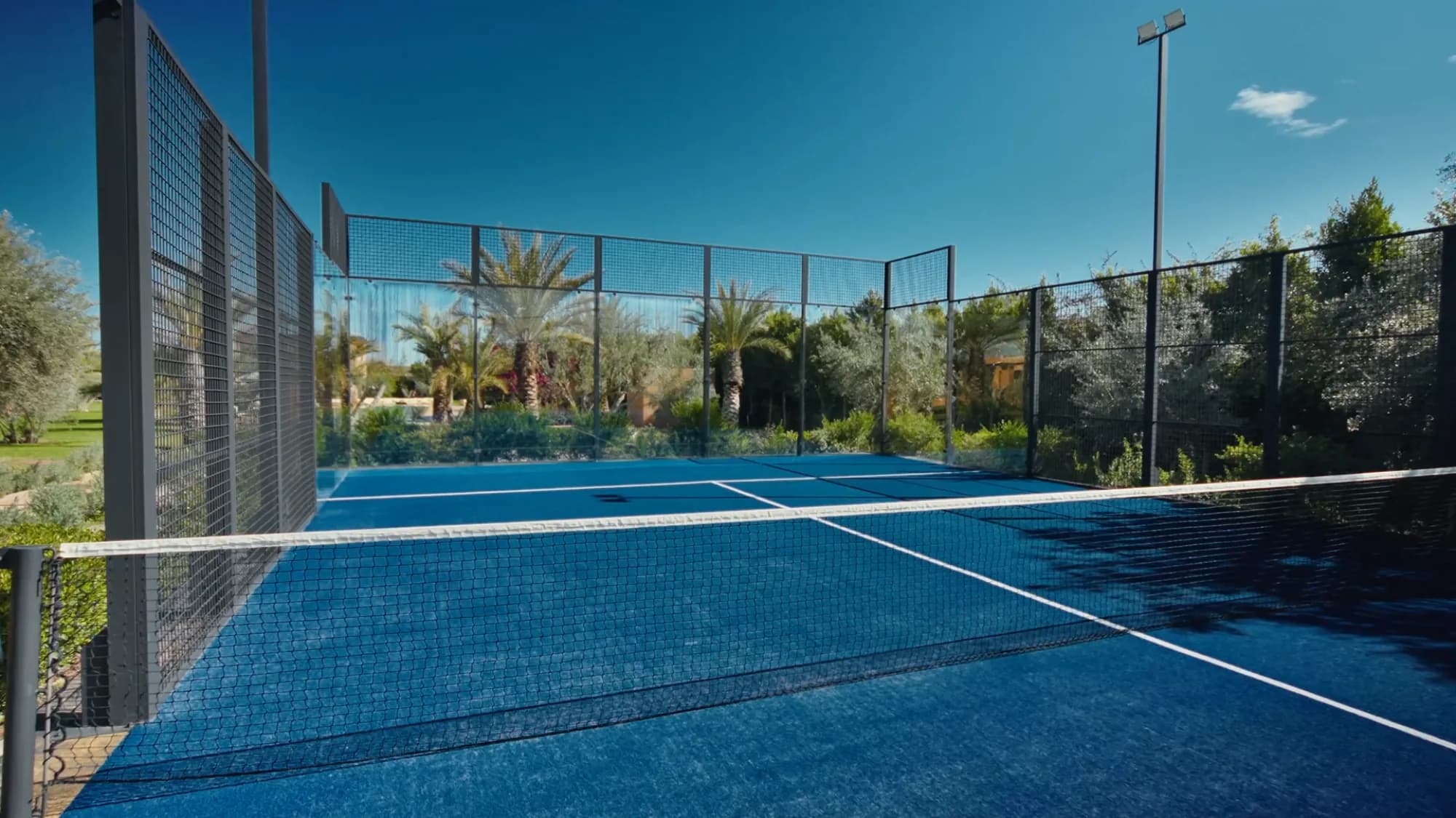 Private padel court at Villa Yemna in Marrakech viewed from inside, with blue playing surface, net and surrounding olive grove.