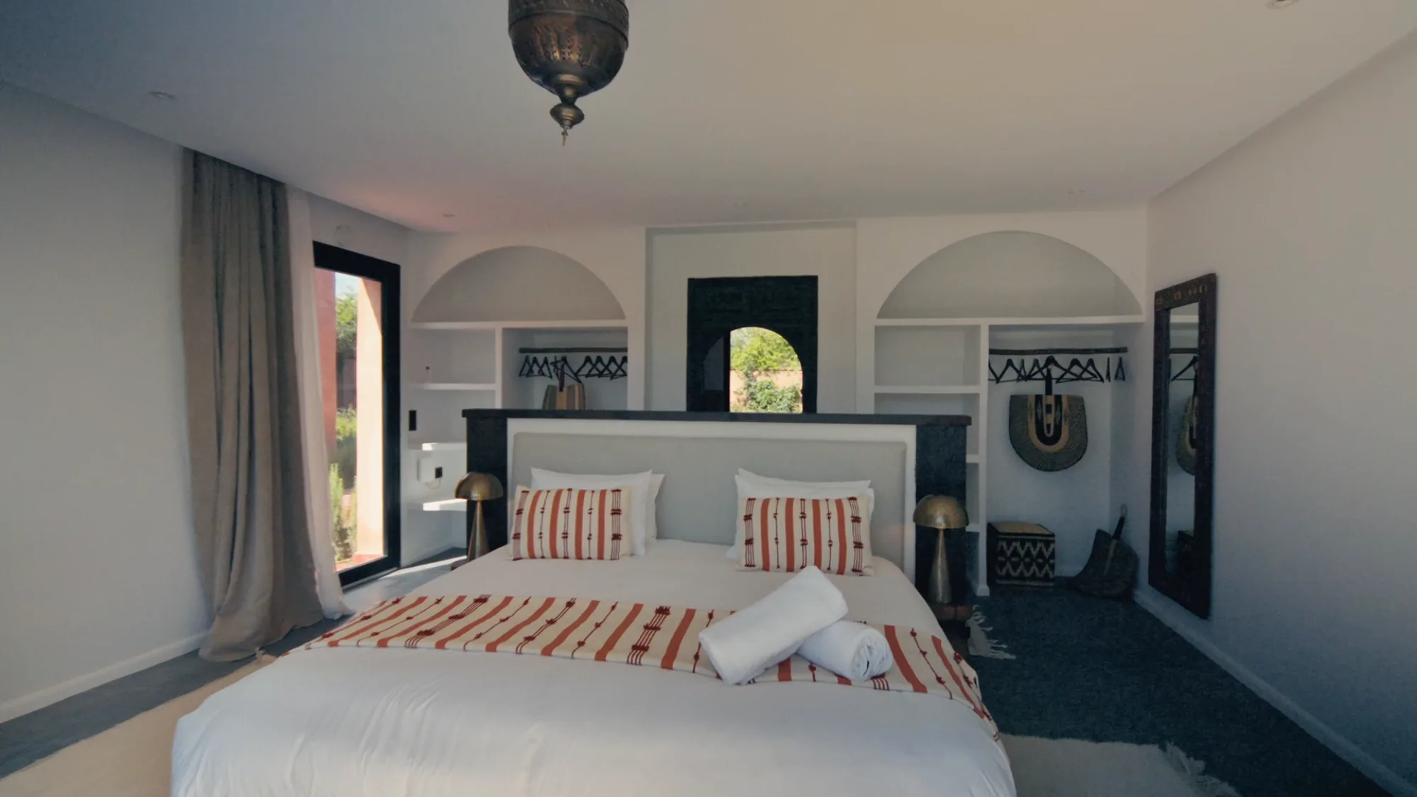 Villa Yemna suite bedroom in Marrakech with king-size bed in white linen, arched niches, terracotta striped pillows and Moroccan pendant light.